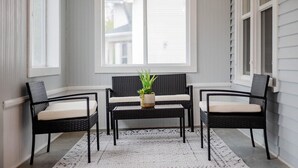 Un coin salon avec un canapé en osier noir et deux fauteuils, une petite table agrémentée d’une plante, et une vue sur une maison par le biais d’une grande fenêtre.