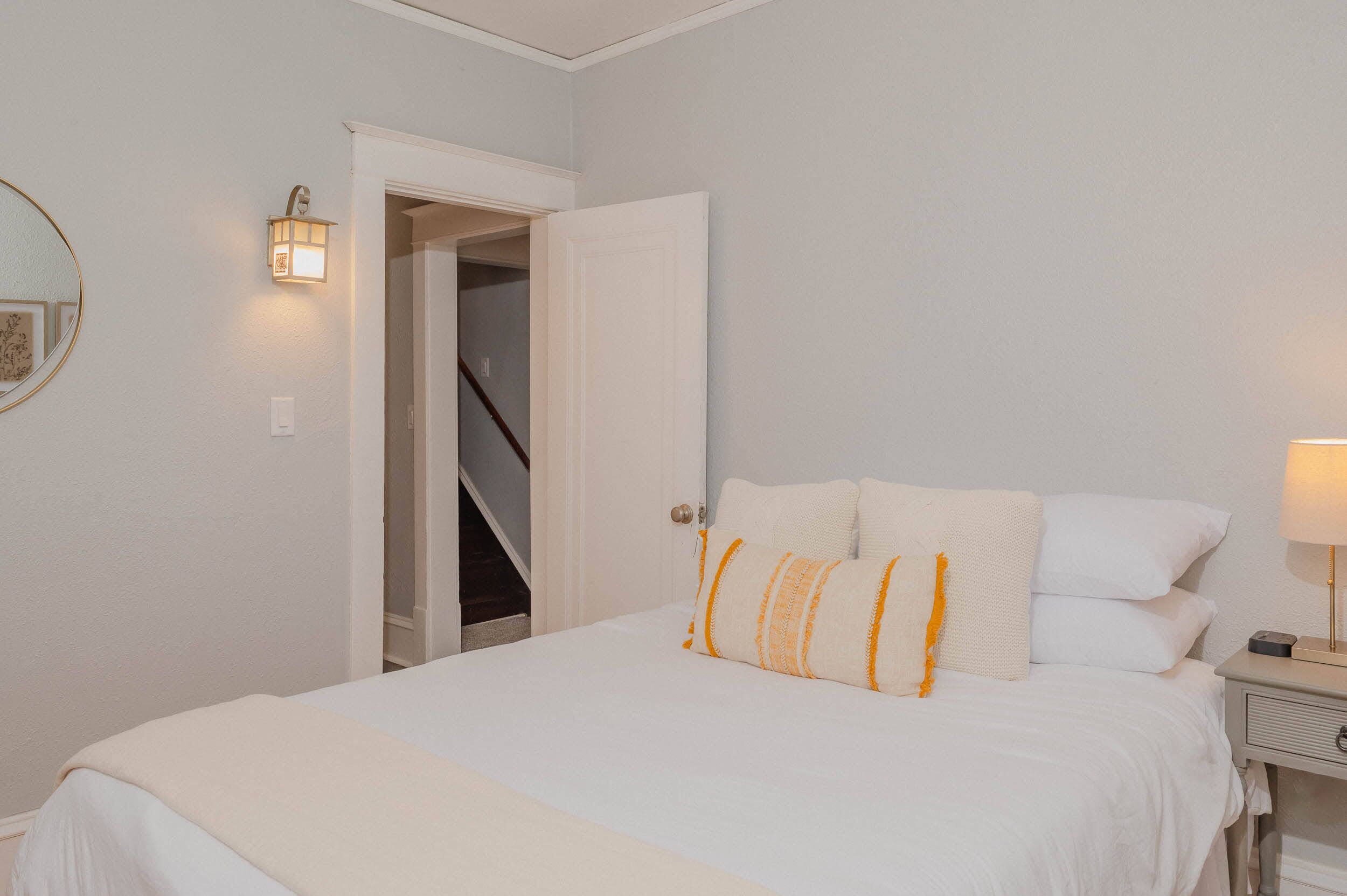 A bedroom with a bed, a nightstand, a lamp, a wall-mounted light, and a door leading to a staircase.