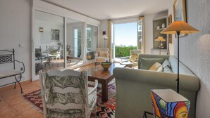 Un salon spacieux doté d’une grande porte-fenêtre coulissante donnant sur un balcon, un coin repas avec une table et des chaises, et un canapé agrémenté d’un coussin coloré.