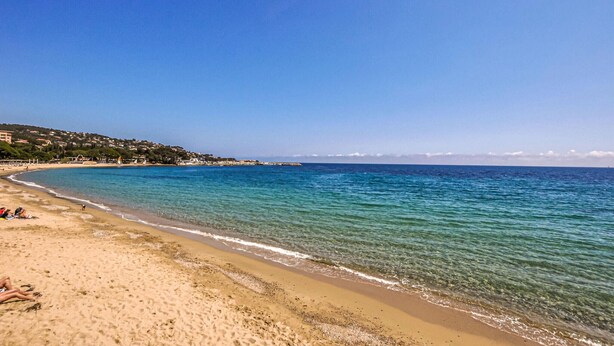 Une plage avec une eau d’un bleu limpide, un rivage sablonneux et des bâtiments en arrière-plan.