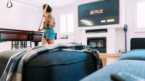 A living room with a television displaying 'NETFLIX', a piano, and a person sweeping. - Glenelg 4BR with pool, 2 min to beach, sleeps 13 (Glenelg South)