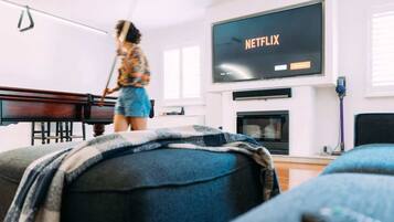 A living room with a television displaying 'NETFLIX', a piano, and a person sweeping.