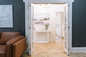 Un pasillo con suelo de madera, un sofá de cuero marrón, una consola blanca con un florero y un cuadro con motivos playeros en la pared.