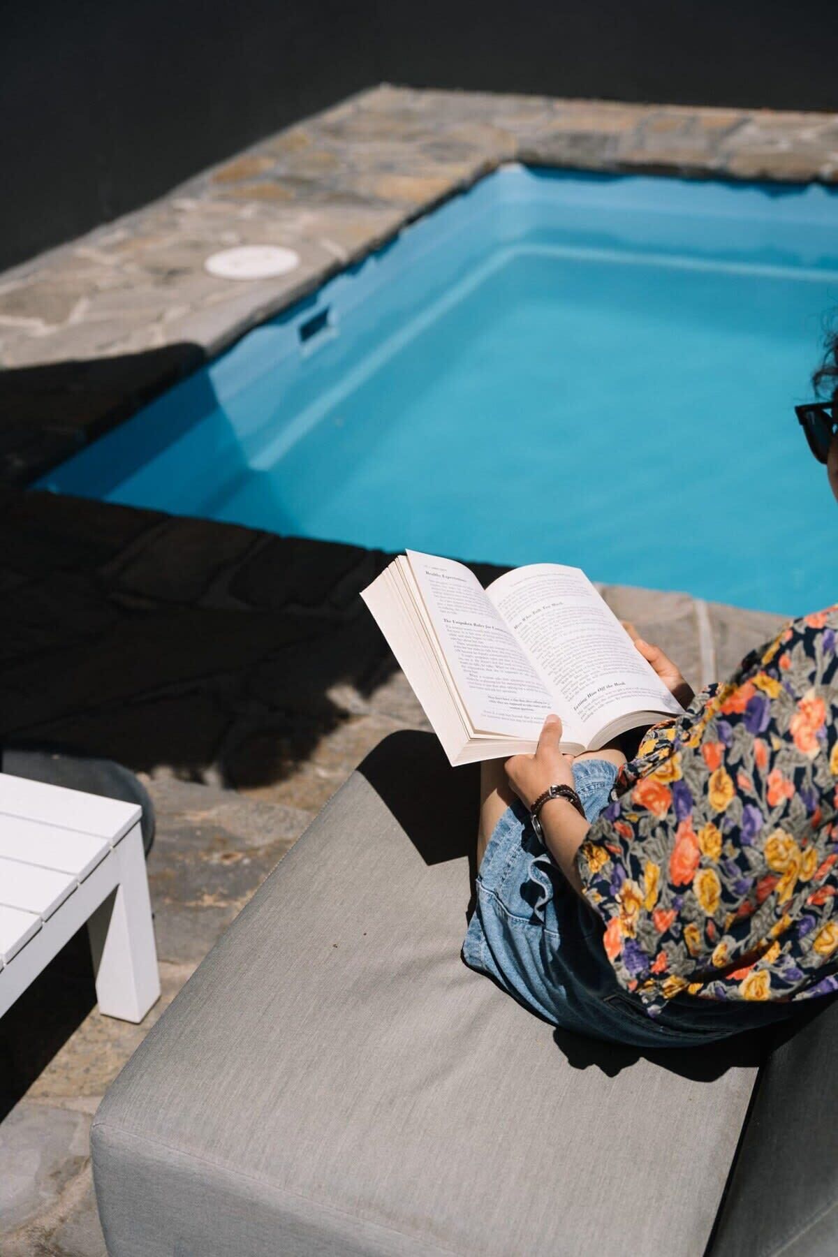 A person reading a book by a pool with a white table and a grey bench.