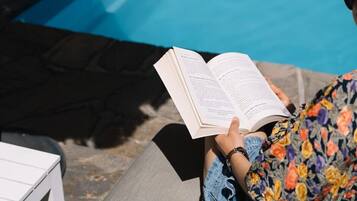 A person reading a book by a pool with a white table and a grey bench.