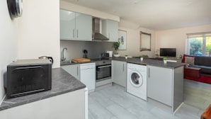 A modern kitchen with a built-in oven, microwave, and dishwasher, a large island with seating, and a dining area with a red sofa. - Bright & Contemporary 1bedroom Annexe - Herne Hill! (London)