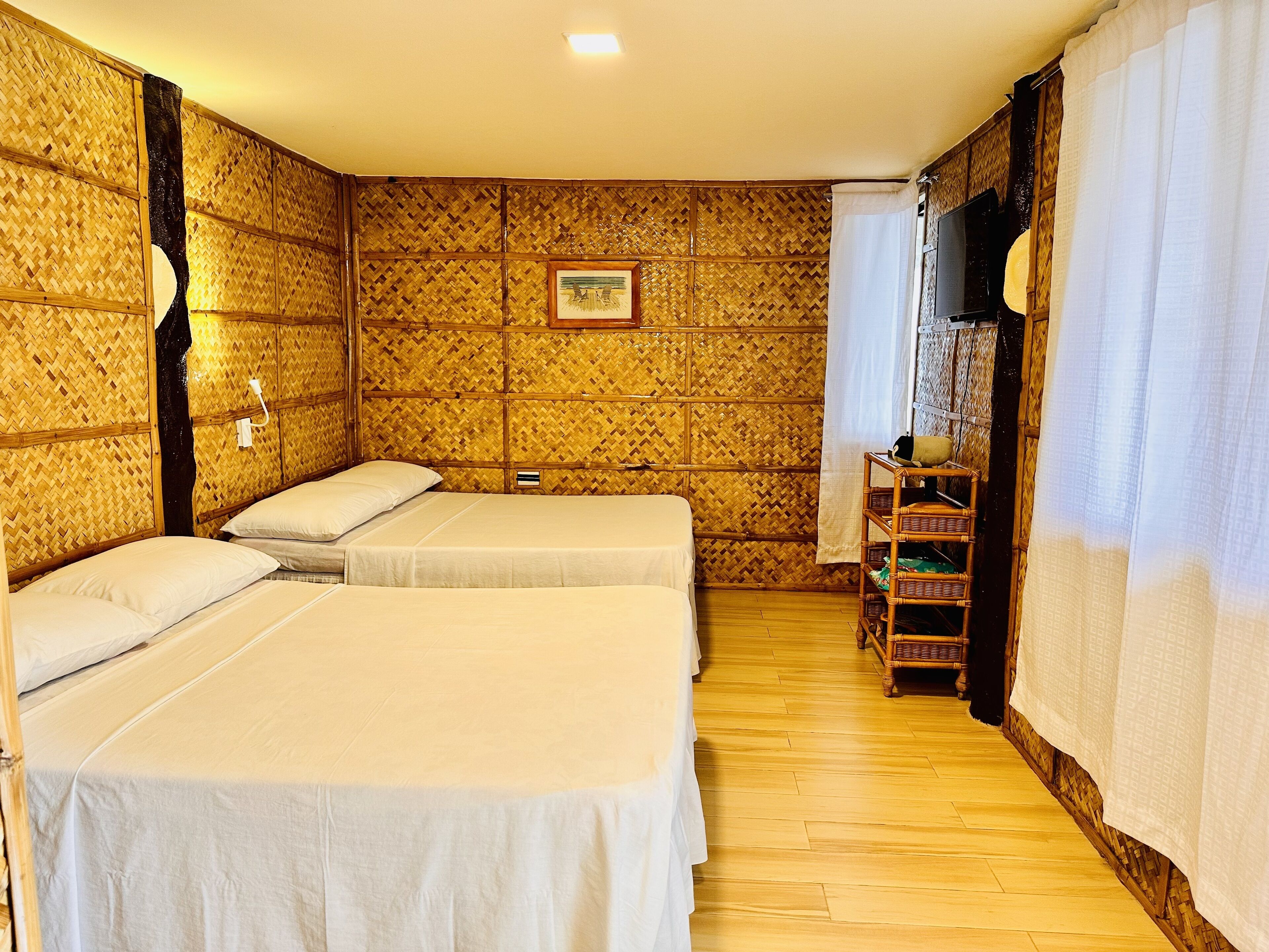 A room with two beds, a wooden floor, a small table with a basket, and a wall with a woven pattern.