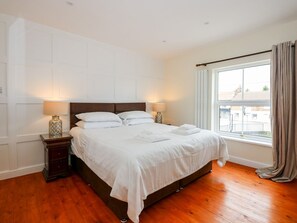 Ein ordentlich bezogenes Bett mit weißen Laken, zwei Nachttische mit Lampen, ein Fenster mit Vorhängen und Holzboden.