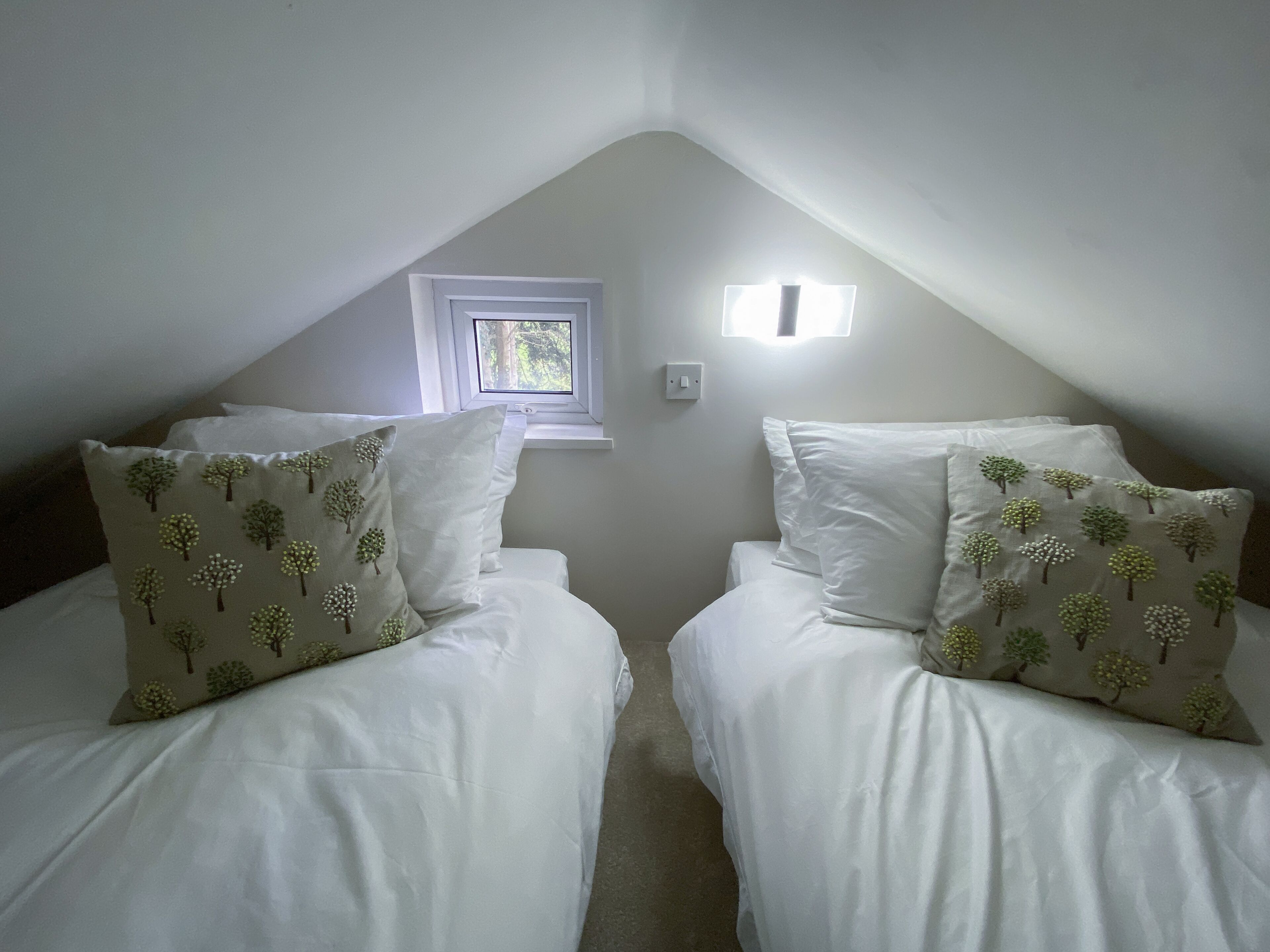 Camera da letto con due letti, ognuno con biancheria bianca e cuscini a motivi floreali, una piccola finestra e un portalampada fissato alla parete.