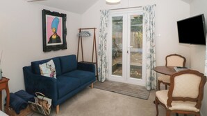A living room with a blue sofa, a wooden chair, a television, and a door leading to a garden. - The Potting Shed (Langport)