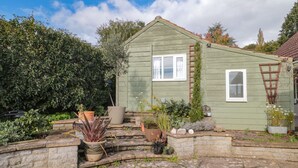 A green wooden house with a stone-paved courtyard, surrounded by various potted plants and a large bush. - The Potting Shed (Langport)