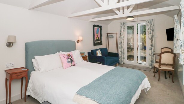 A bedroom with a bed, bedside tables, a chair, a sofa, a TV, and a balcony with curtains. - The Potting Shed (Langport)