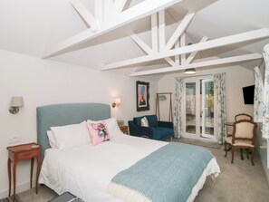 A bedroom with a bed, bedside tables, a chair, a sofa, a TV, and a balcony with curtains. - The Potting Shed (Langport)