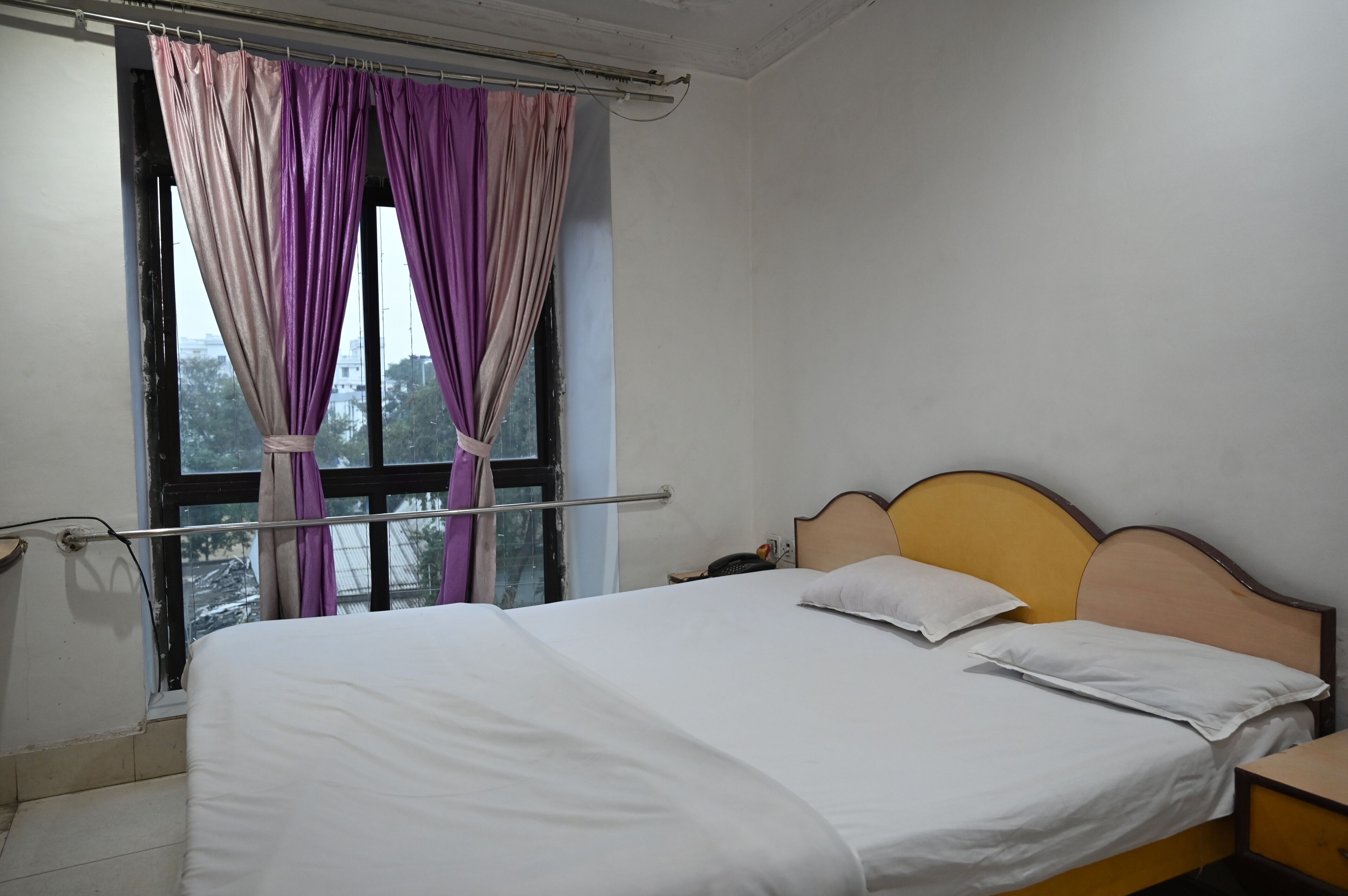 A single bed with a wooden headboard, a bedside table, and a window with purple curtains.