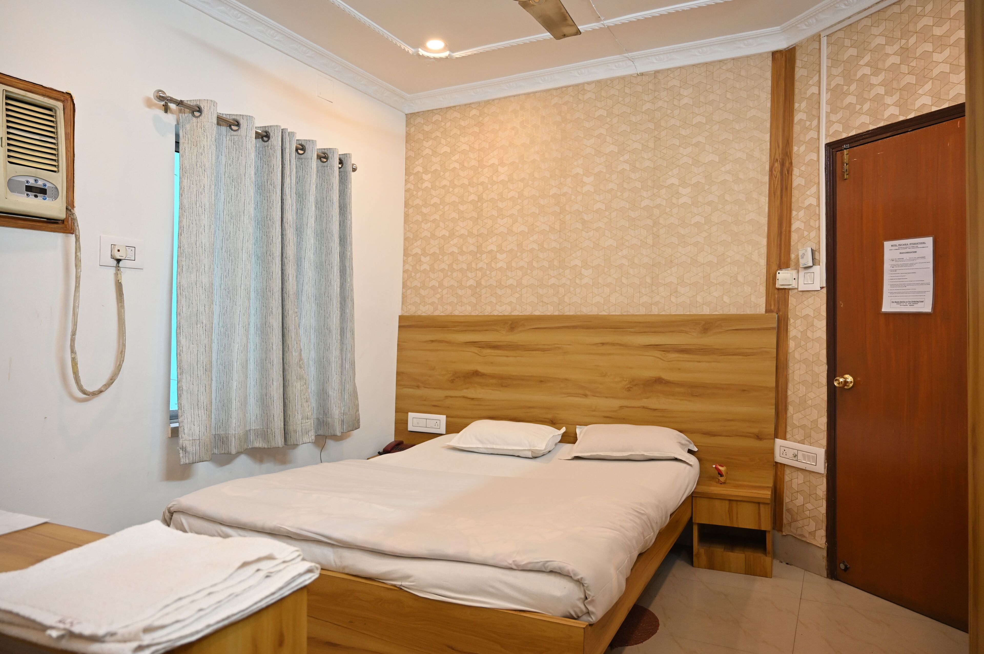 Un dormitorio con cabecera de madera, cama con ropa blanca, mesita de noche y tocador de madera, radio montada en la pared y una puerta con un cartel.