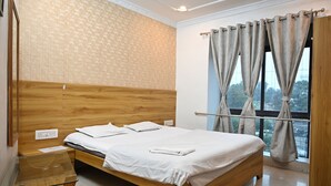 Quarto de hotel com cabeceira de madeira, mesa lateral de vidro, cama com roupa de cama branca e janela com cortinas.