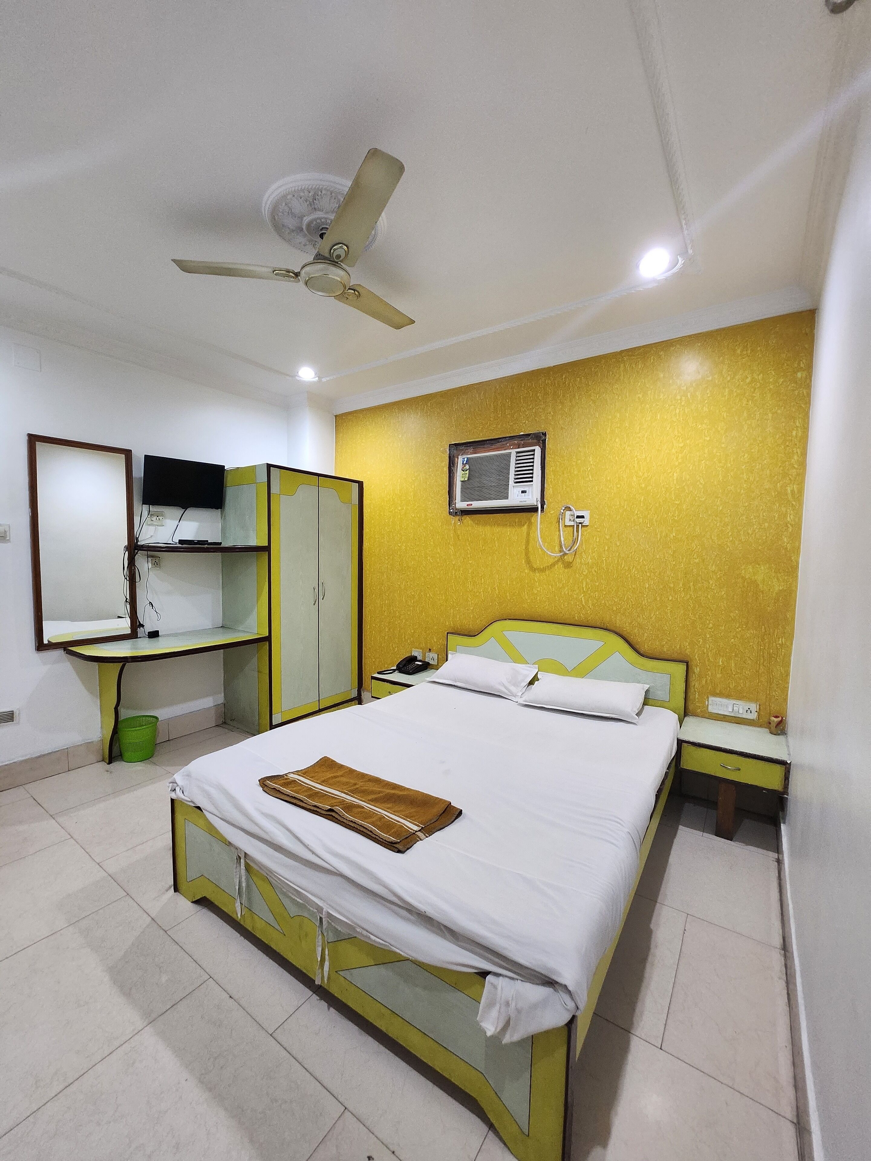 A hotel room with a bed, a desk, a wardrobe, a ceiling fan, an air conditioning unit, and a television.