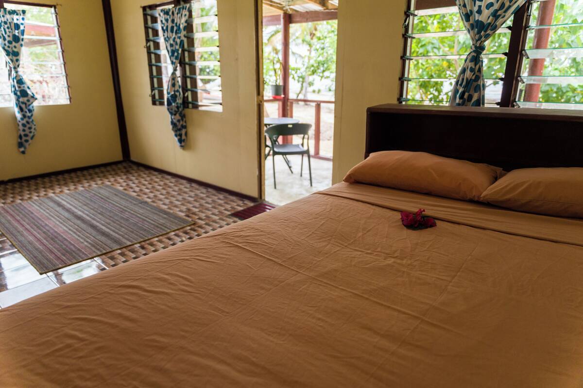 Un dormitorio con cama, ventana con cortinas, una silla cerca y vista a zonas verdes.
