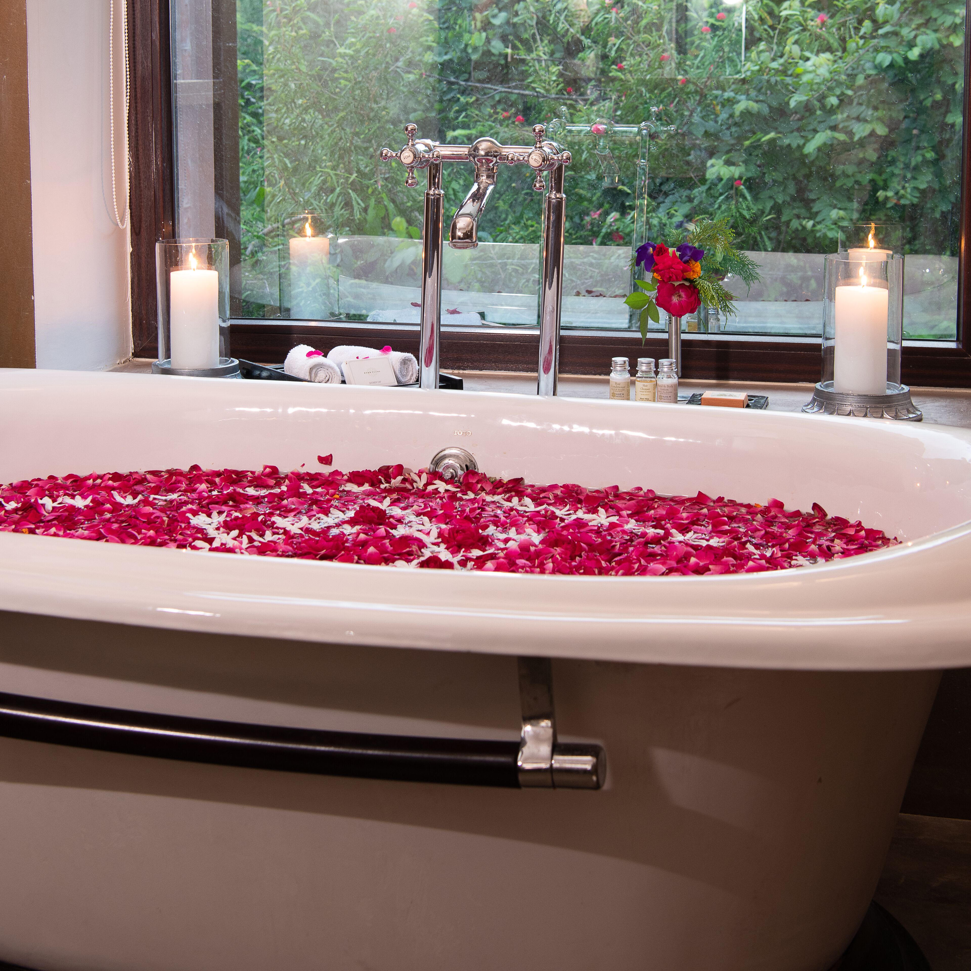 A bathtub filled with rose petals, a window with a view of greenery, and two lit candles.
