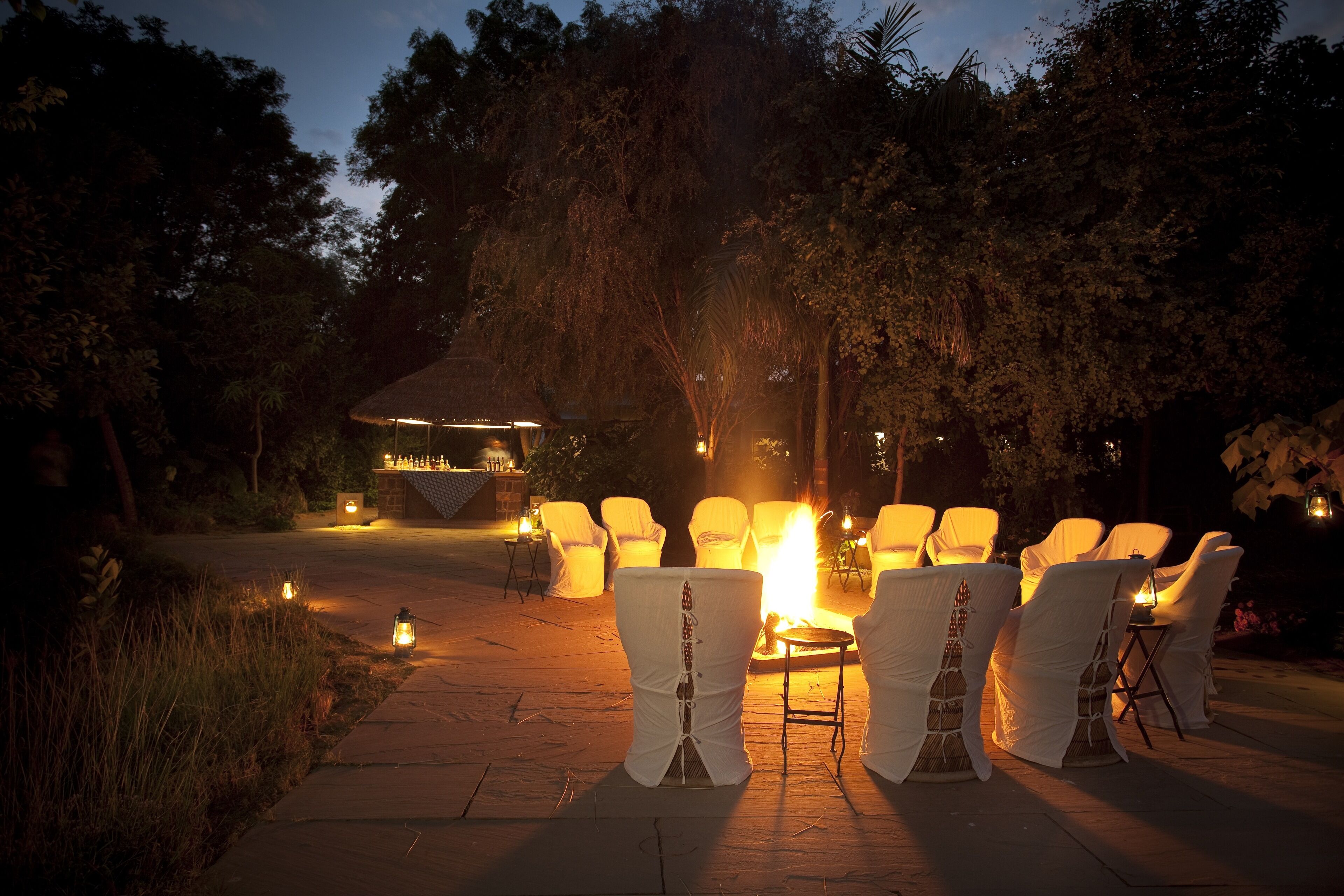 Un espace extérieur aménagé avec des chaises et des tables blanches, entouré d’arbres et agrémenté d’une petite structure illuminée.
