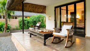 Un patio couvert avec une table à manger, des chaises et un fauteuil à bascule. Il est surmonté d’une structure à toit de chaume et offre une vue sur le jardin.