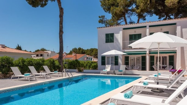 Una piscina con sillones de playa y sombrillas, rodeada por un edificio con techo de tejas y contraventanas verdes.