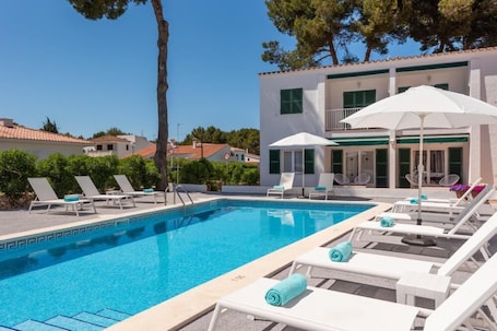 A swimming pool with lounge chairs and umbrellas, surrounded by a building with a tiled roof and green shutters.