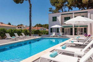 Una piscina con sillones de playa y sombrillas, rodeada por un edificio con techo de tejas y contraventanas verdes.
