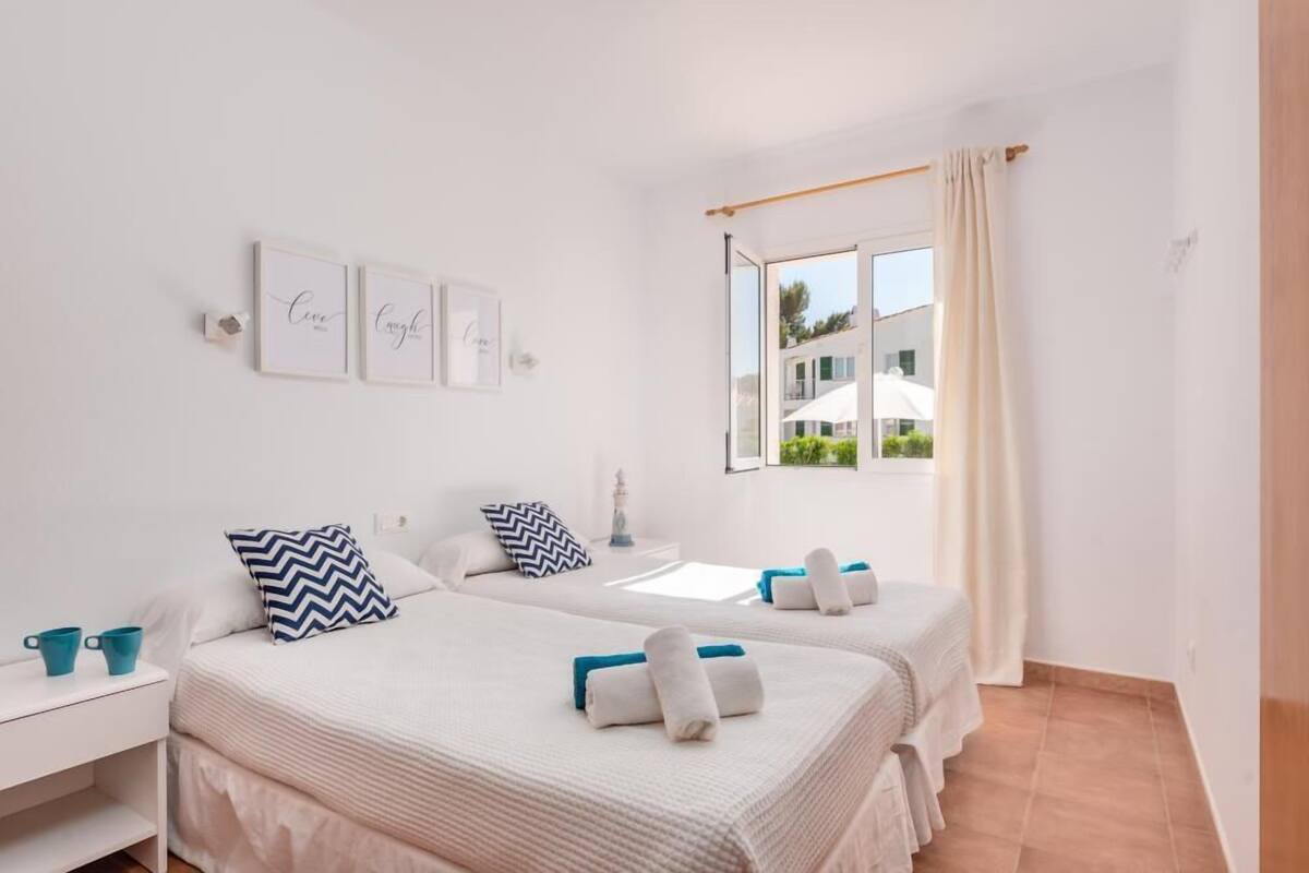 Une chambre d’hôtel avec deux lits, une fenêtre donnant sur des immeubles et un mur blanc orné de tableaux.
