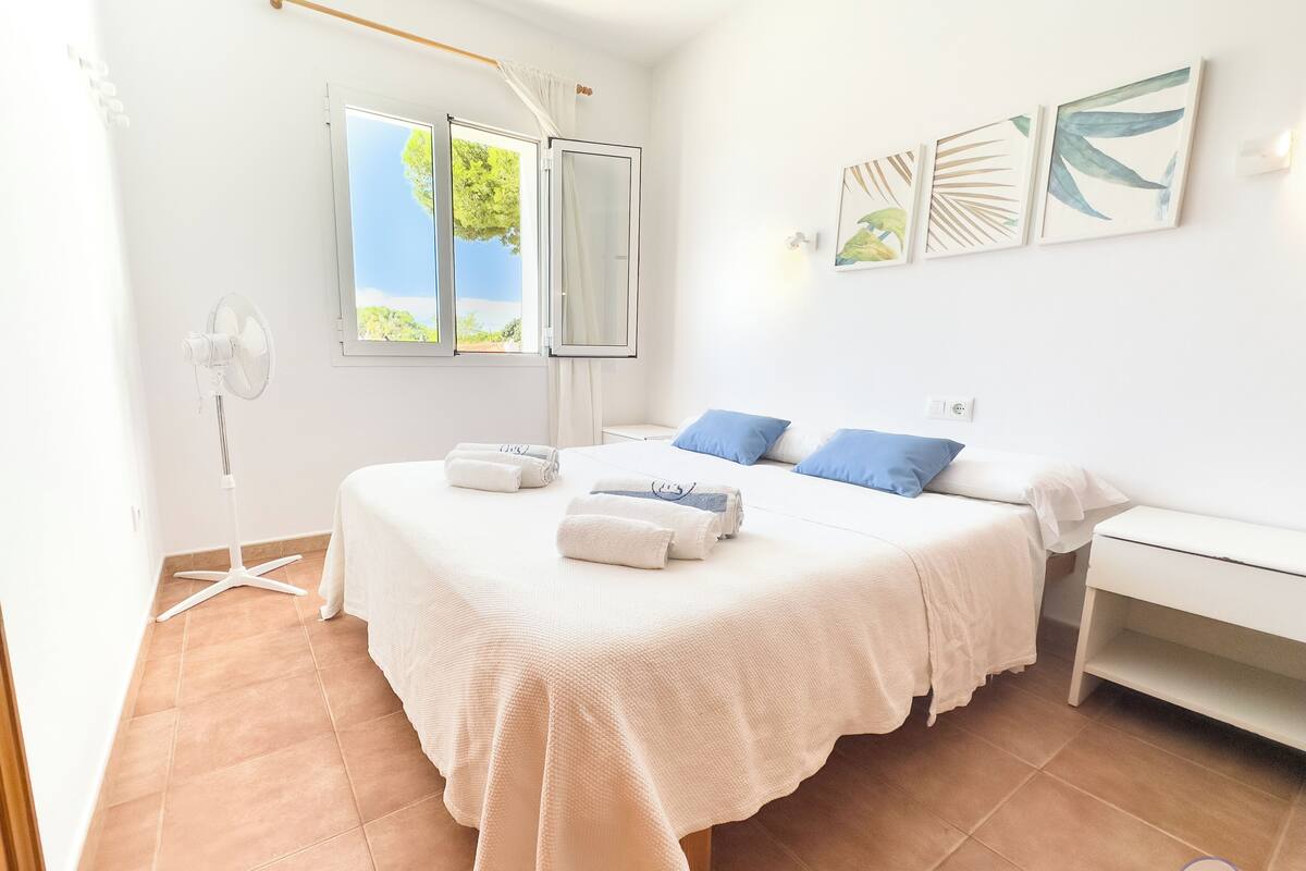 Une chambre à coucher avec un lit, deux tables de chevet, un ventilateur sur pied et une fenêtre avec des rideaux.