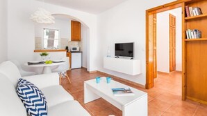 Una sala de estar moderna con un sofá blanco, una mesa de centro y un televisor. Hay una zona de cocina con un microondas y una pequeña mesa de comedor.