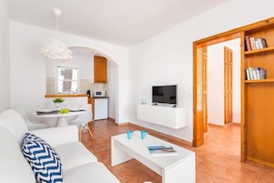 Una sala de estar moderna con un sofá blanco, una mesa de centro y un televisor. Hay una zona de cocina con un microondas y una pequeña mesa de comedor.