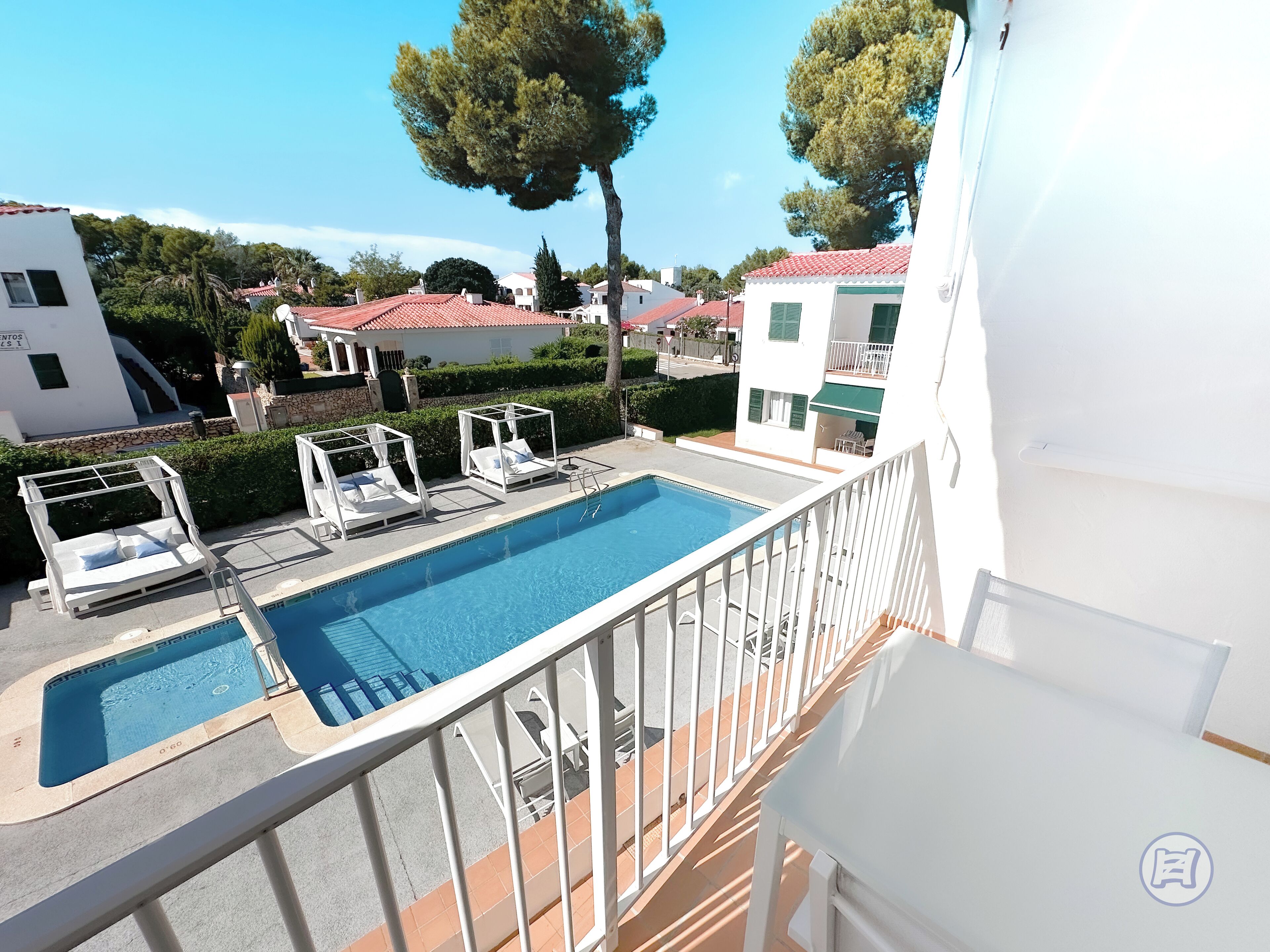 A swimming pool with lounge chairs, a white building with a red roof, and a tree in the background.
