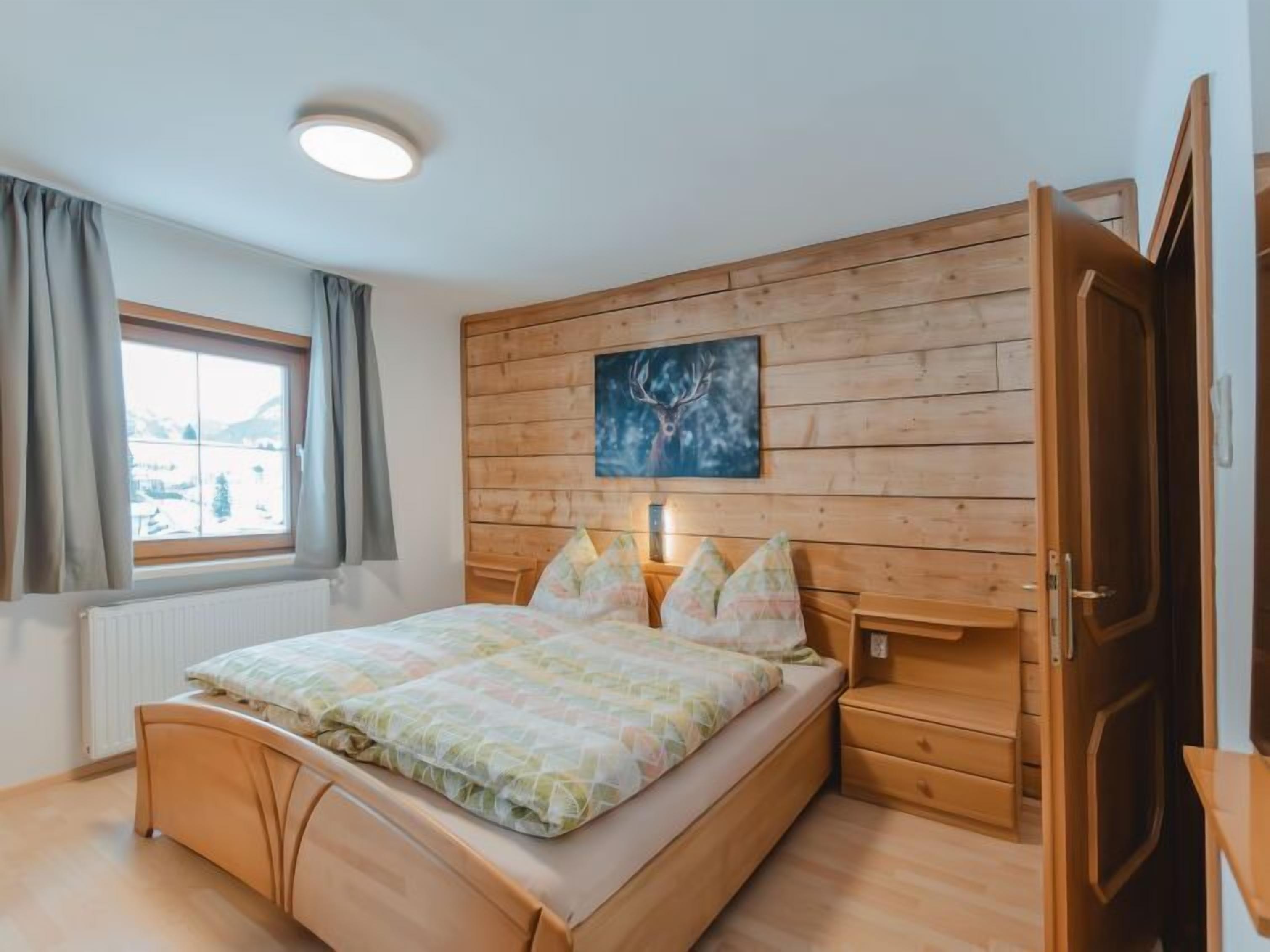 Une chambre à coucher avec une tête de lit en bois, un lit recouvert d’une couvre-lit à motifs floraux, une fenêtre avec des rideaux et un tableau représentant un cerf.