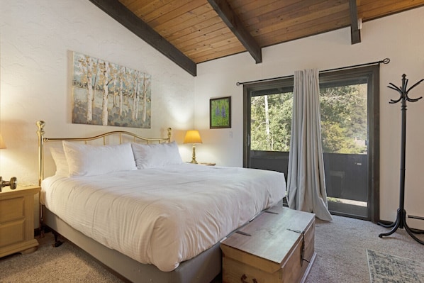 A bedroom with a large bed, wooden headboard, two bedside tables with lamps, a window with curtains, and a view of trees outside. - West Vail Ptarmigan Townhome - Hot Tub & Easy Bus (Vail)