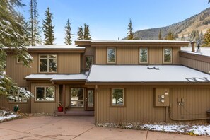 A two-story house with a snow-covered roof, a front porch, and a view of a forested mountain.