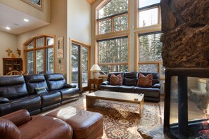 A spacious living room with leather sofas, a coffee table, and large windows offering a view of trees.