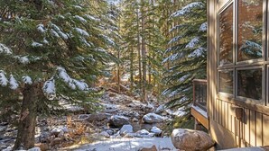Eine Holzhütte mit einer Holzterrasse, umgeben von schneebedeckten Bäumen und einem steinigen Bach.