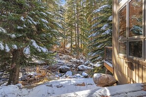 A cabin with a wooden deck, surrounded by snow-covered trees and a rocky stream. - Large East Vail Home - Steps to Bus & Gore Creek (Vail)