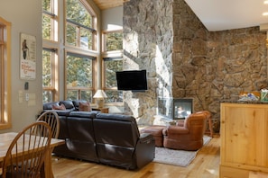 A cozy living room with a stone wall, large windows, a fireplace, and a flat-screen TV.