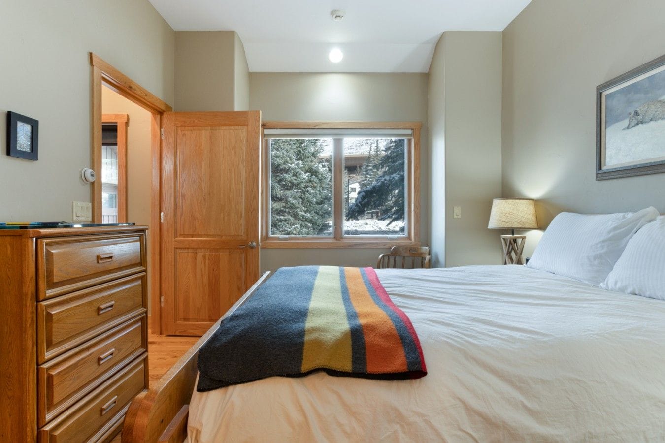 A bedroom with a bed, a wooden dresser, a window with a view of trees, and a lamp.