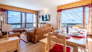 Una habitación acogedora con una mesa de madera preparada para comer, un sofá y un televisor. Amplias ventanas con vistas a la montaña.