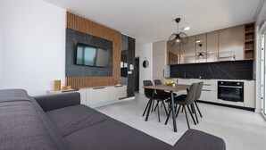 A modern kitchen with a built-in oven, a dining area with a wooden table and chairs, and a large TV mounted on a wall with a wooden panel design. - Apartments Twins with pools (Novalja)