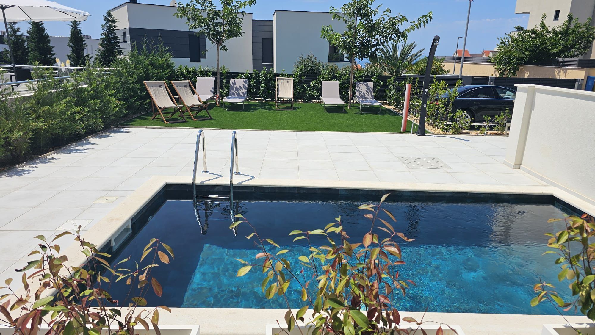 A swimming pool with a metal ladder, surrounded by a tiled patio with lounge chairs and a small grassy area.