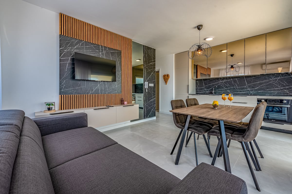 A modern living room with a large sectional sofa, a dining table with chairs, and a kitchen area with a large island and a built-in oven.
