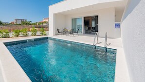 A modern pool area with a clear blue pool, a covered seating area with chairs, and a glass door leading to a building. - Apartments Twins with pools (Novalja)