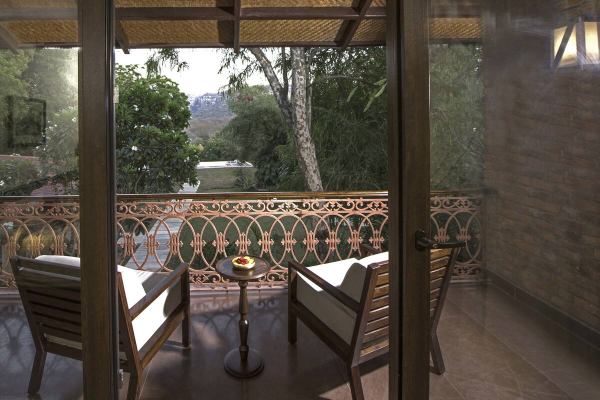 Un balcon donnant sur un jardin et des arbres. On y trouve deux chaises en bois et une petite table ronde sur laquelle est posé un saladier rempli de fruits.