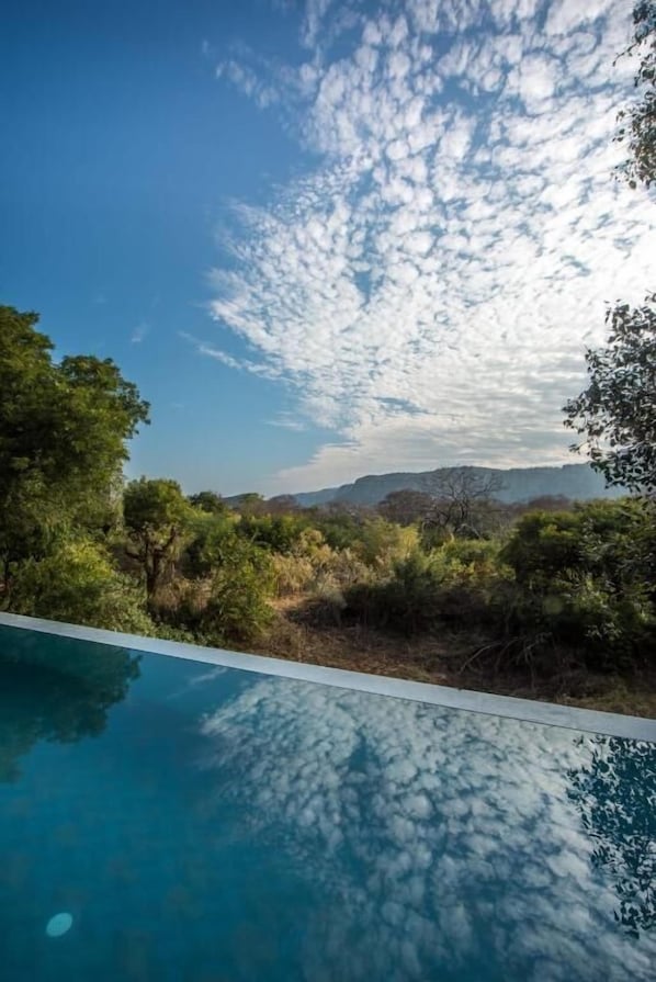 Un ciel d’un bleu limpide parsemé de quelques nuages, une piscine qui reflète le ciel et une vue sur un paysage montagneux à la végétation luxuriante.