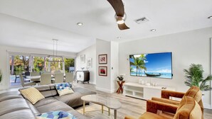 A modern living room with a large sofa, a coffee table, and a TV mounted on the wall. There is a dining area with a table and chairs visible through an open door. - Lakefront Family Fun Home with Private Dock 3BR 2B (Fort Lauderdale)