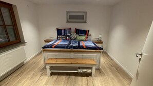 A bedroom with a wooden floor, a bed with a blue and orange bedspread, and a small bedside table with a lamp. - Kellerwohnung (Seelbach)